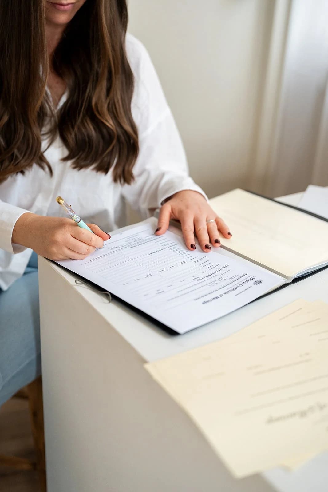 A close-up of hands signing the official marriage certificate at a desk, with paperwork laid out beside it.