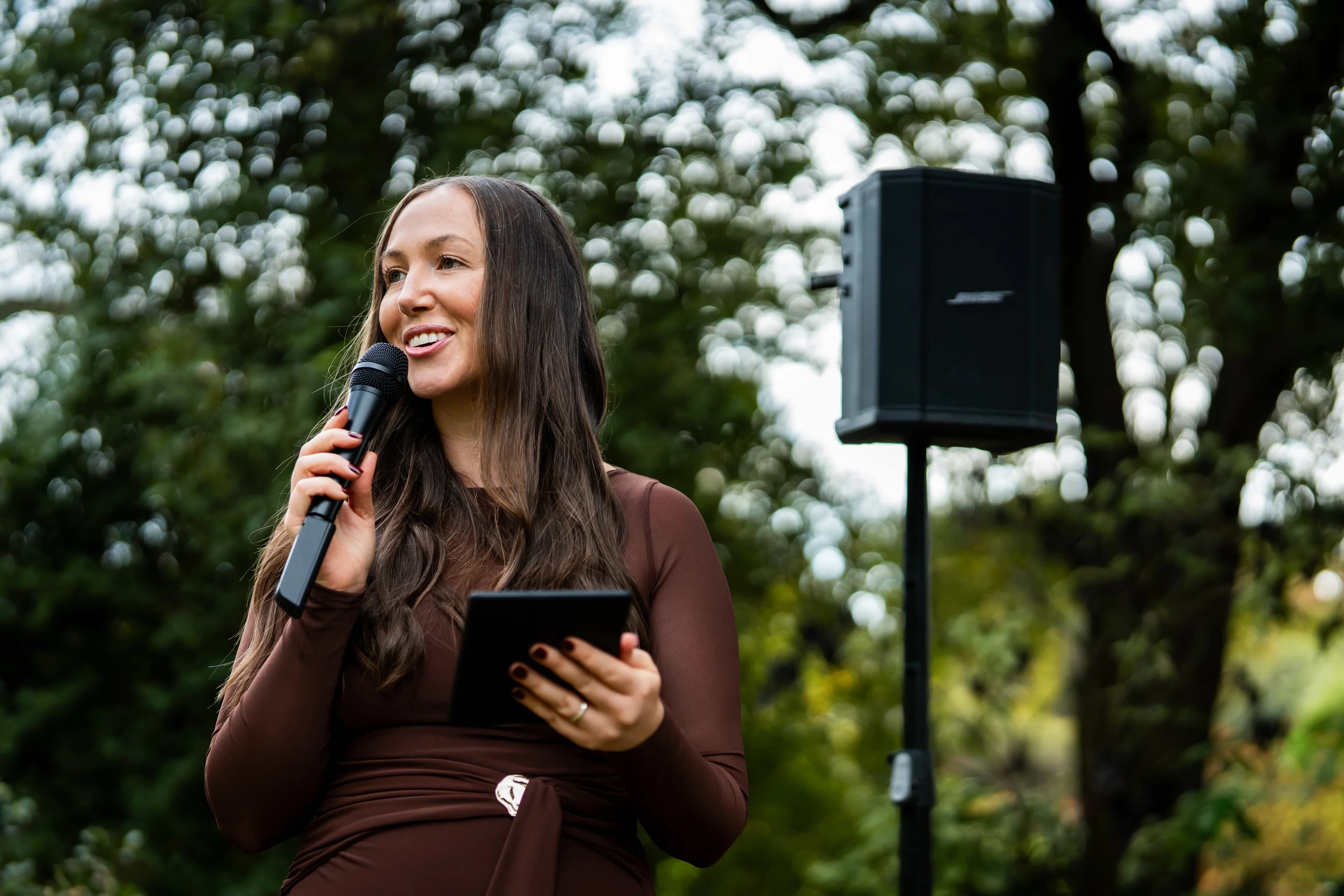 Tam speaking into a microphone, with her PA speaker on a stand beside her, in a leafy garden.