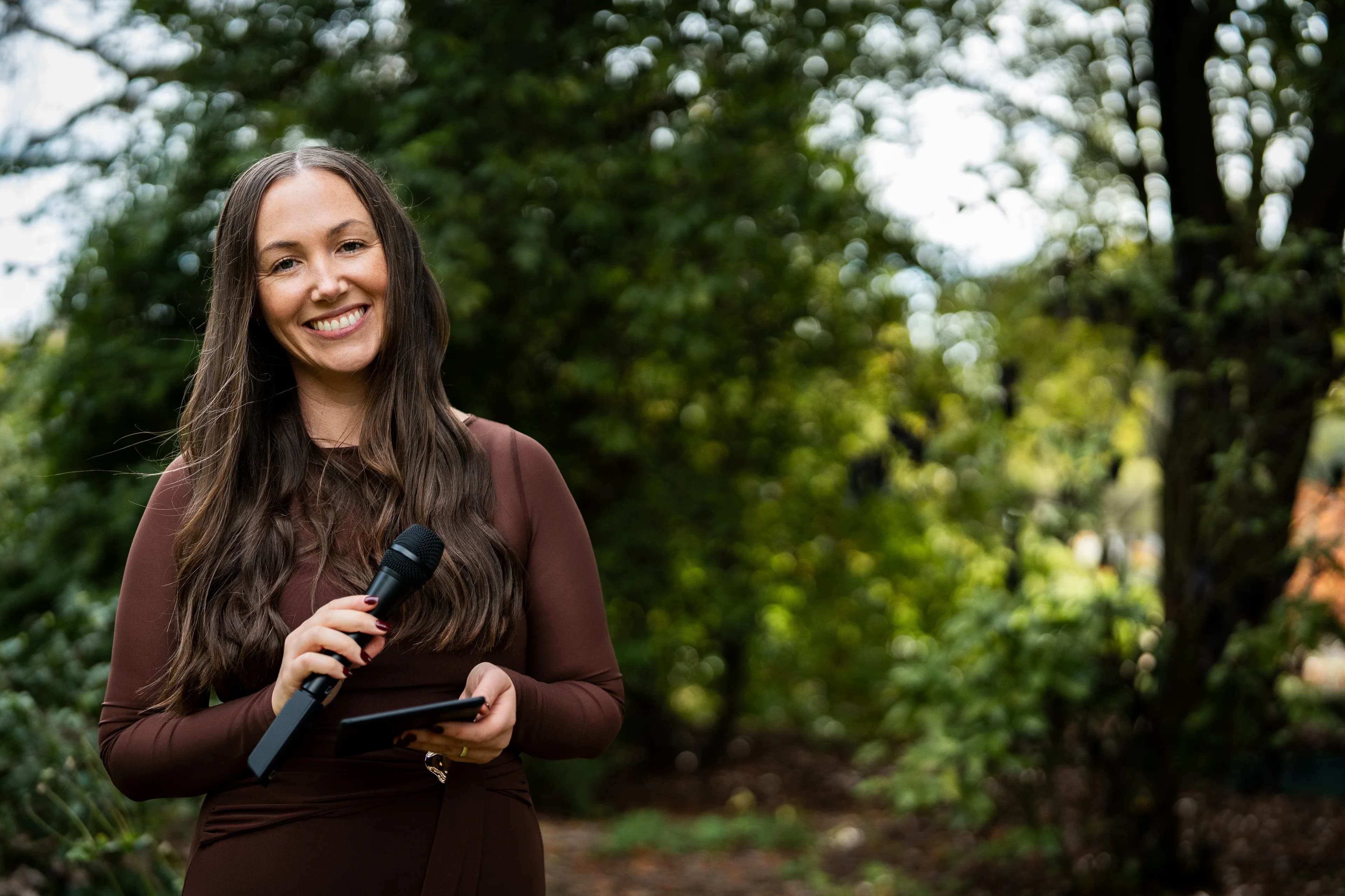 Tam smiling, holding a microphone and her ceremony notes in a leafy garden.