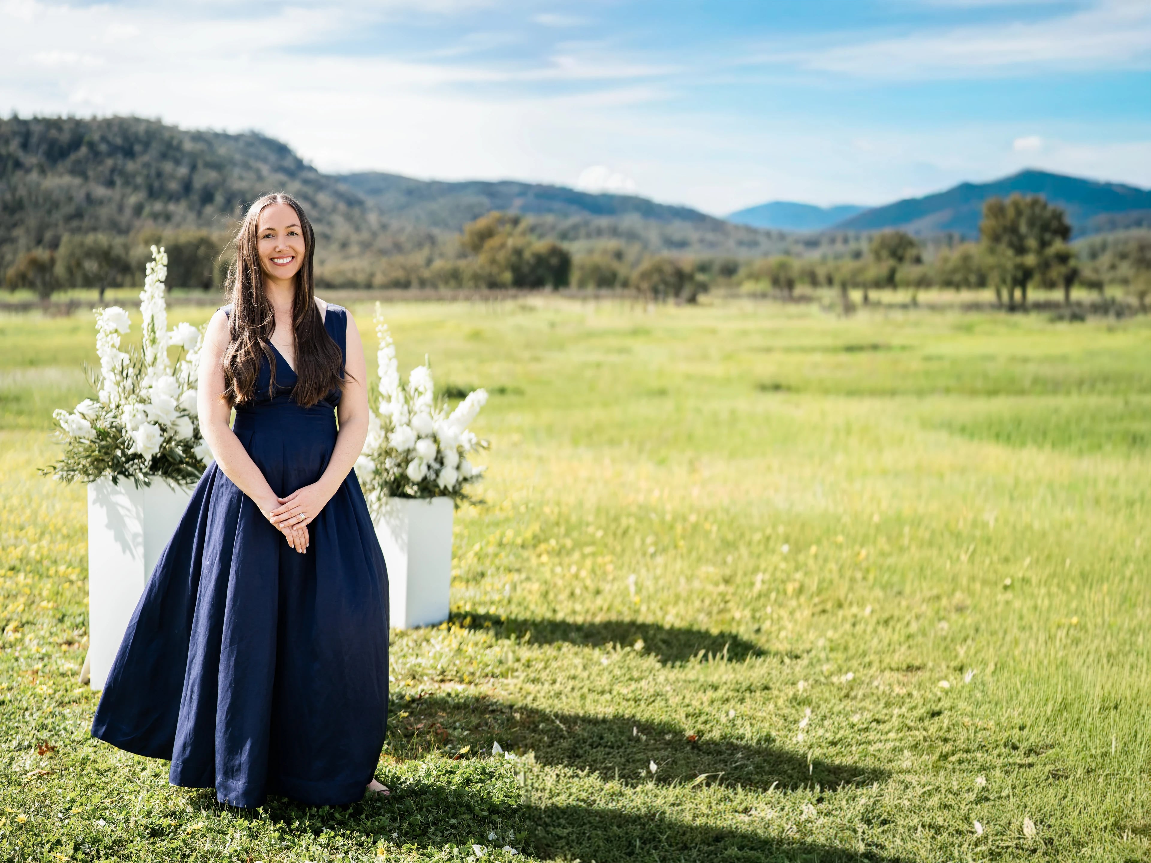 Tam, smiling in a navy gown beside two arrangements of white flowers in a sunlit green field with hills behind.