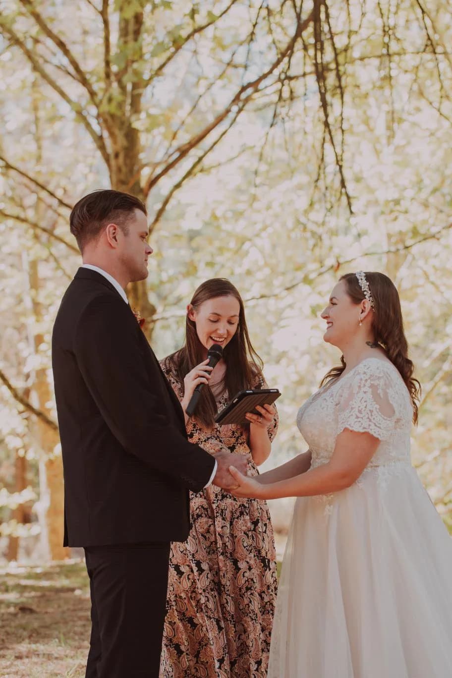 Tam leading a couple's outdoor ceremony, holding a microphone as they hold hands beneath golden autumn trees.