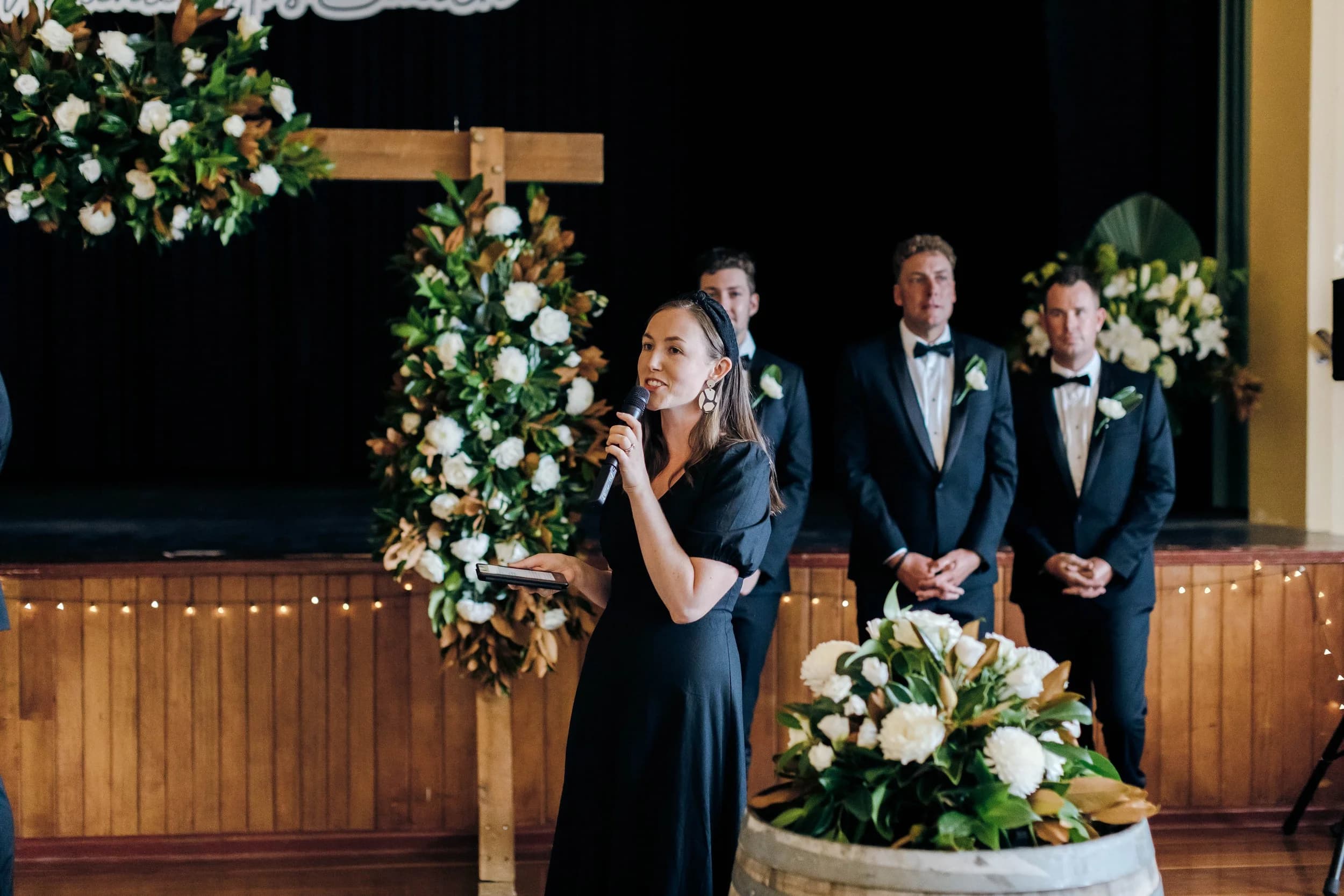 Tam speaking into a microphone as she leads a wedding ceremony in a hall, with three groomsmen standing behind her.