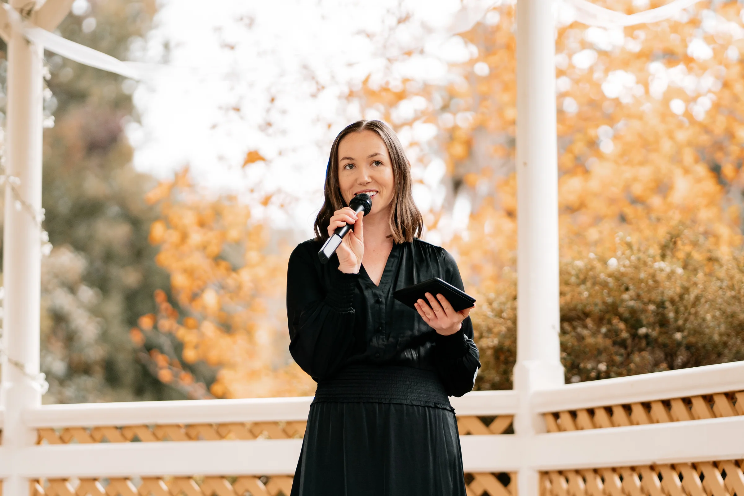 Tam speaking into a microphone and holding a tablet under a white pergola, autumn-coloured trees behind her.