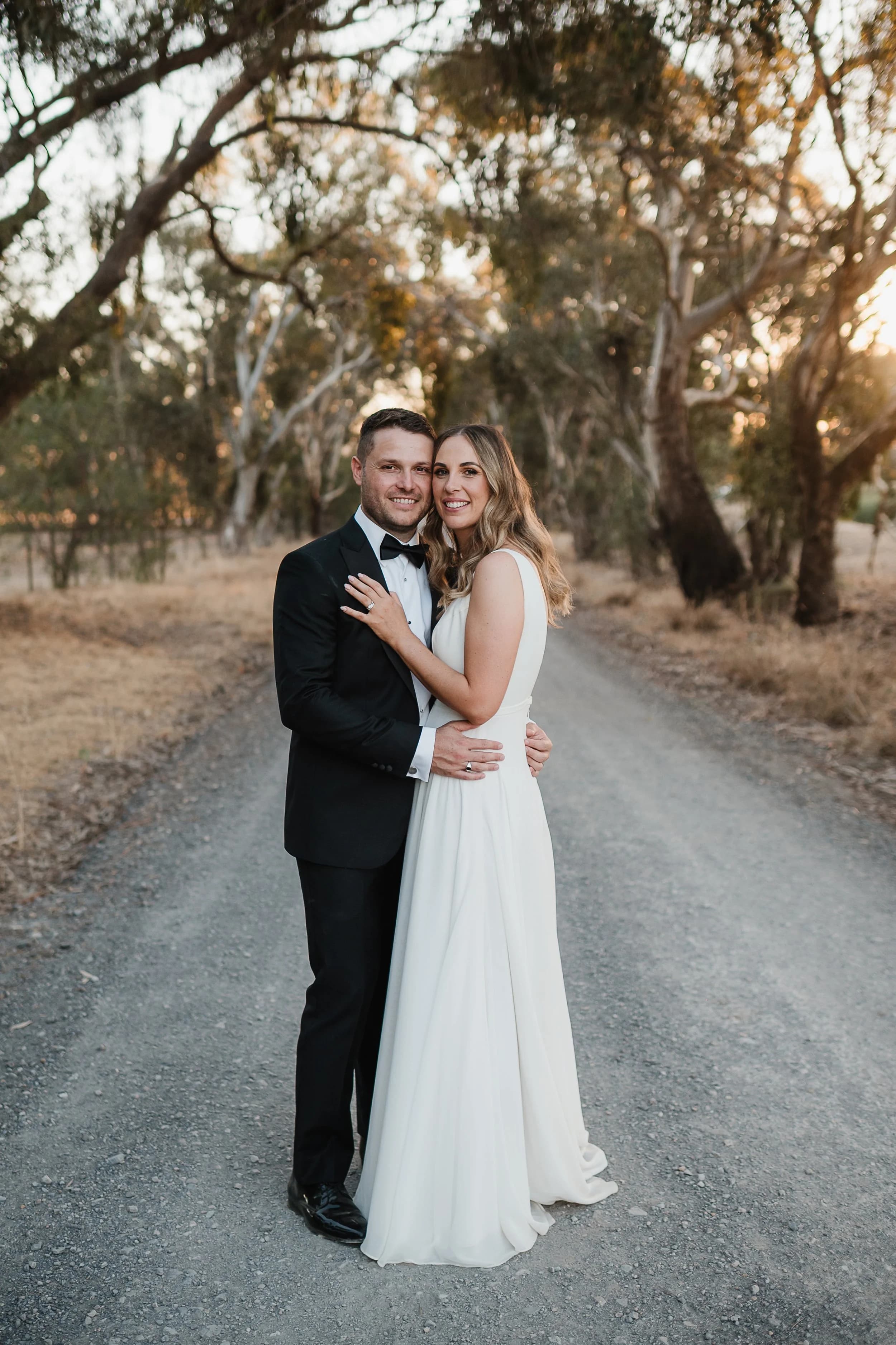 Rhiannon and Sam smiling in each other's arms on a gum-lined gravel country road at golden hour.