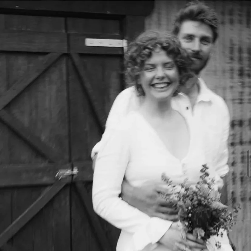 Em and Hame laughing together beside a timber barn door, in black and white.